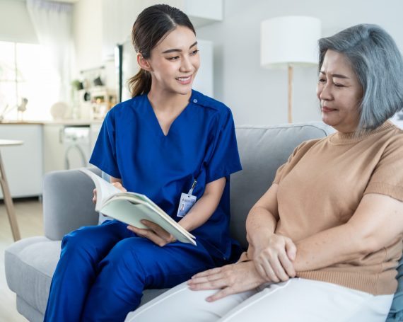 Asian woman nurse taking care of senior elderly female at nursing home. Young Caregiver Therapist doctor sit on sofa, read a book to happy older handicapped patient. Medical insurance service concept.