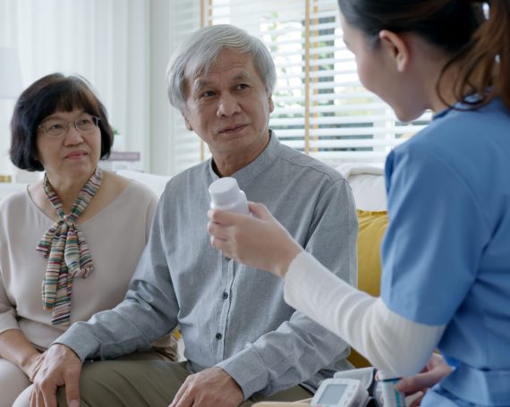 Young caregiver in scrubs uniform showing medicine bottle to elderly asian couple man and woman in home visit care nursing service.  Asian senior with assisted living medication monitoring concept.