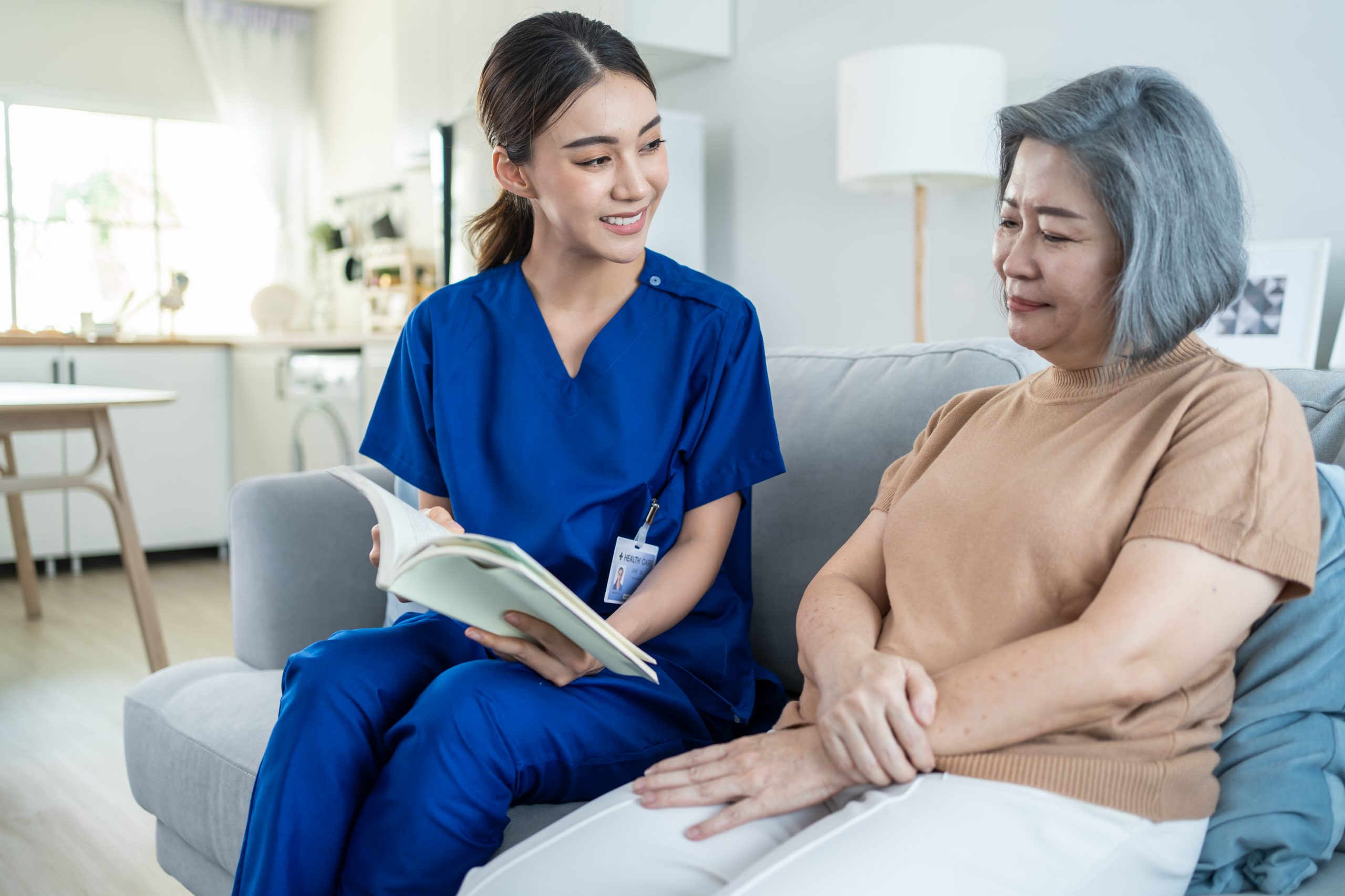 Asian woman nurse taking care of senior elderly female at nursing home. Young Caregiver Therapist doctor sit on sofa, read a book to happy older handicapped patient. Medical insurance service concept.