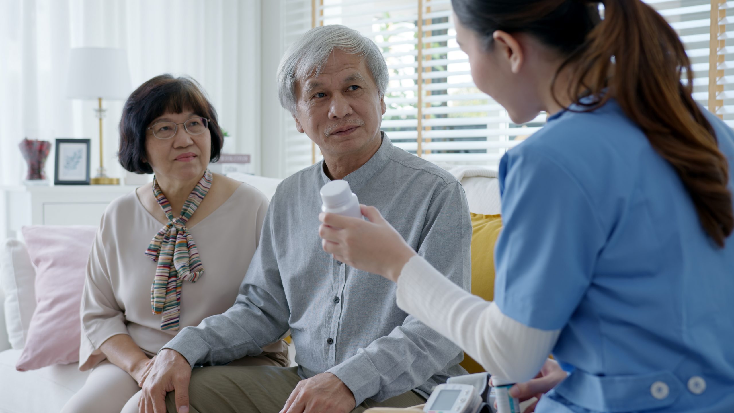 Young caregiver in scrubs uniform showing medicine bottle to elderly asian couple man and woman in home visit care nursing service.  Asian senior with assisted living medication monitoring concept.